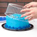A hand placing a blue cake into a D&W Fine Pack plastic cake container on a table in a bakery display.