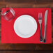 A white plate with a glass and silverware on a red Hoffmaster scalloped paper placemat.