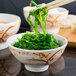 A close up of chopsticks holding green seaweed in a round gold bowl.
