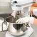 A person wearing gloves uses a KitchenAid wire whip to mix dough in a white mixer.