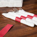 A wood table with a stack of white napkins with a red rectangular band around a group of forks.