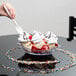 A hand holding a white spoon over a banana split in a clear dish.