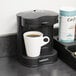 A black Conair Cuisinart coffee maker on a counter next to a white mug of coffee.