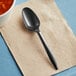 A black Choice medium weight plastic teaspoon on a napkin next to a bowl of food.