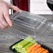 A hand holding a Fineline clear plastic snack tray dome lid over celery and carrots.