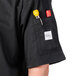 A close-up of a Mercer Culinary black chef coat pocket with a pen and yellow and silver objects.