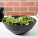 A black 10 Strawberry Street stoneware serving bowl with salad on a table.