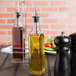 A Choice oil and vinegar cruet with a pourer on a table with a bottle of oil and a pepper shaker.