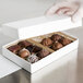 A hand holding a 7" x 4 3/8" white candy box filled with brown chocolates.