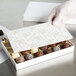 A hand in a white glove placing a 9 1/4" x 5 1/2" white candy box with gold floral pattern on a table.
