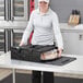 A woman holding a black Choice insulated deli tray and party platter bag full of food in front of a toaster oven.
