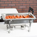 A Vollrath stainless steel chafing dish with meatballs in it on a table with other food and a tray of sauce.