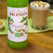 A green bottle of Monin pistachio syrup next to a cup of coffee.