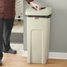 A man in a chef's uniform standing next to a Rubbermaid beige square commercial trash can with a lid that opens with a foot pedal.