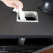 A hand using a white tissue to put a white bag into a rectangular stainless steel in-counter trash chute.