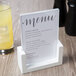 A white Cal-Mil U-frame tabletop card holder displaying a menu on a table next to a glass of liquid.