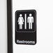 A black and white 9" x 6" restroom sign with man and woman pictograms.