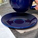 A hand holding a Tuxton cobalt blue cappuccino cup over a saucer on a blue table.