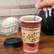 A hand using a Royal Paper black beverage plug and stirrer in a cup of coffee on a counter.