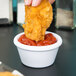 A person holding a fried chicken nugget and dipping it into a white Carlisle ramekin of ketchup.