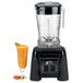 A Waring commercial blender with a glass of orange juice next to it.