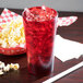 A red Cambro plastic tumbler filled with ice and a red drink on a table with a bowl of popcorn.