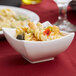 A white square porcelain bowl filled with pasta, vegetables, and olives.