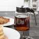 A Tablecraft glass syrup dispenser with a black lid on a table with pancakes and syrup.