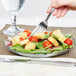 A hand holding a Carlisle Petal Mist clear polycarbonate plate with a fork over a salad with tomatoes and croutons.