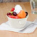 A bowl of fruit with whipped cream in a ivory melamine bouillon cup with a fork.