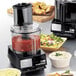A Waring food processor with various food in the bowl on a table.