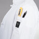 A person wearing a white Chef Revival chef coat with a pen and a thermometer in the pocket.