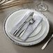 A white Charge It by Jay round charger plate with a silver band on the rim with silverware and a napkin on it.