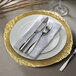 A white charger plate with a gold leaf embossed rim with silverware and a white napkin.