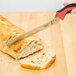 A Mercer Culinary Millennia Colors bread knife with a red handle cutting a piece of bread on a cutting board.