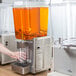 A woman pours orange juice from a Crathco refrigerated beverage dispenser into a clear glass.