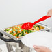 A person holding a red Cambro salad bar spoon over a pan of vegetables.
