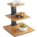 A Cal-Mil bamboo 3 tier riser with bamboo shelves holding bowls, cups, and fruit on a wooden table.