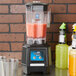 A Waring Torq 2.0 Blender on a counter with a glass of orange juice and bottles of liquid.