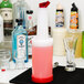 A Carlisle white plastic bottle with a red cap and spout on a bar counter full of pink liquid.