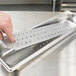 A person's hand holding a Vollrath stainless steel false bottom over a metal tray.