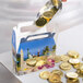 A hand using a metal scoop to pour gold coins into a 1/2 lb. Landscape Window Candy Box on a table.