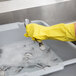 A hand wearing a yellow glove washing a Vollrath gray high density polyethylene bus tub filled with spoons.
