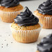 A group of white fluted baking cups filled with cupcakes with black frosting on a counter.