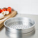 An American Metalcraft aluminum pizza pan with holes on the sides sitting on a table next to tomatoes and garlic.