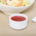 A white Carlisle ramekin filled with red sauce next to a bowl of salad with vegetables.
