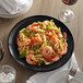 A Tuxton Concentrix black china plate with a fork and a plate of pasta with shrimp and basil on a table.
