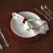 A Tabletop Classics by Walco red plastic charger plate on a table with a white napkin and silverware.