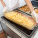 A person in white gloves holding a Cambro Amber High Heat Plastic Food Pan filled with french fries.