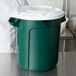 A man in a white shirt standing next to a green Rubbermaid BRUTE recycling can with a white lid.